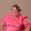 A photograph of Alison Bergblom Johnson, a white woman with short, asymmetrical blonde hair wearing a shiny, pink shirt in front of a light tan background.