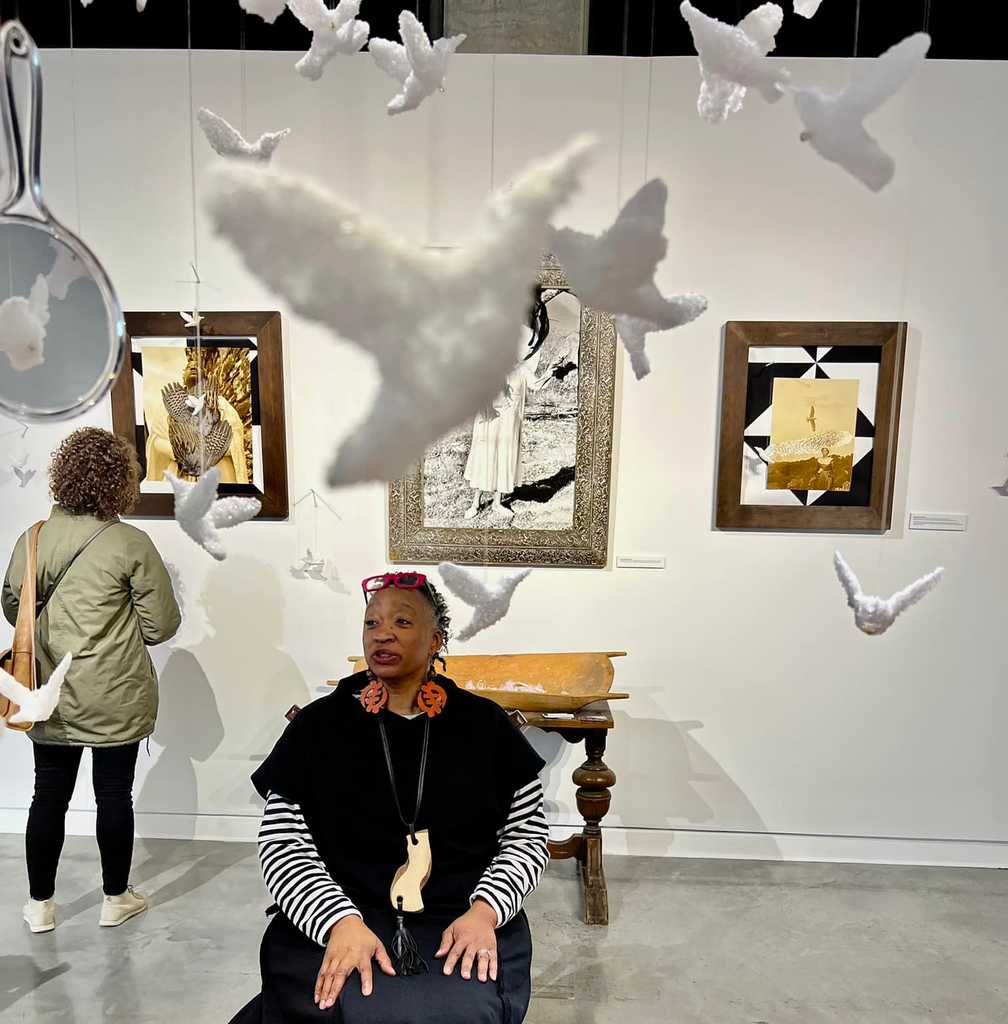 Image description: Hawona Sullivan Janzen, a Black woman wearing a black sweater and skirt sits in her show at the Katherine Nash gallery, several sculpted birds and collages are also visible.