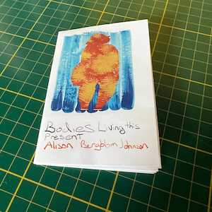 A silhouette of a fat figure with her hair in a short bob cut into yellow and orange marbled paper. The silhouette is on a blue painted background. Below the collage is the zine title handwritten: 'Bodies Living this Present' and Alison Bergblom Johnson's name. The zine rests on a green cutting mat with yellow markings.
